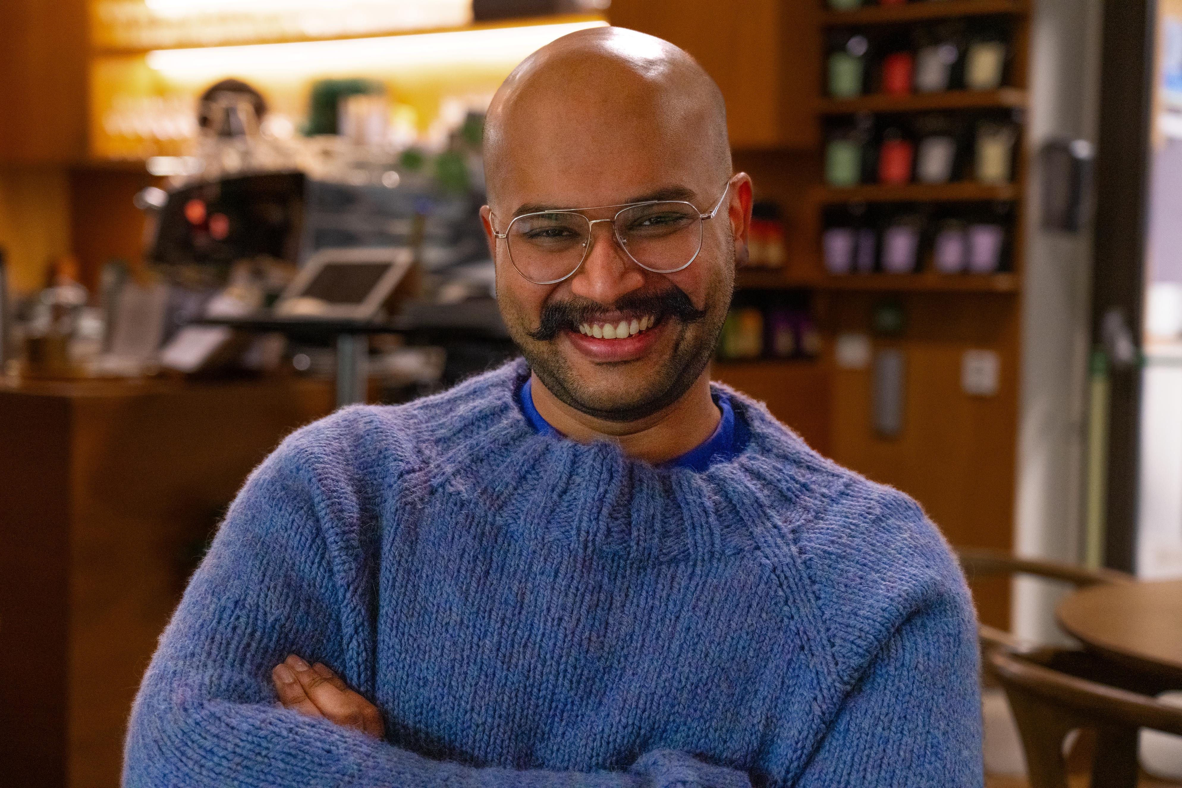 A man wearing a blue sweater, smiling inside of a coffee-shop. There are books in shelves behind him.