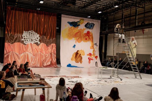 A group of seated people watching a performance. The performers are sitting and standing in a ladder. There's a huge painting in the background