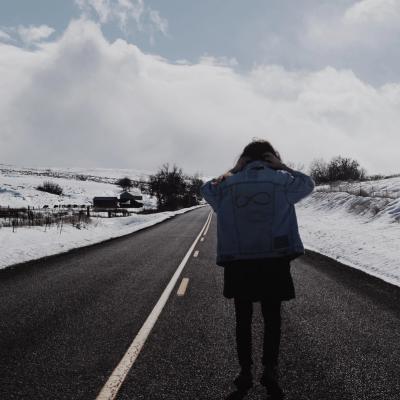 A longhaired man wearing a denim-jacket standing in the the middle of the road with his back turned. There are snow on the sides of the road, and a big cloudy sky
