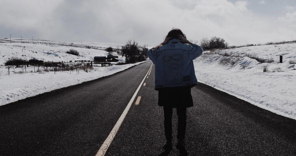 A longhaired man wearing a denim-jacket standing in the the middle of the road with his back turned. There are snow on the sides of the road, and a big cloudy sky