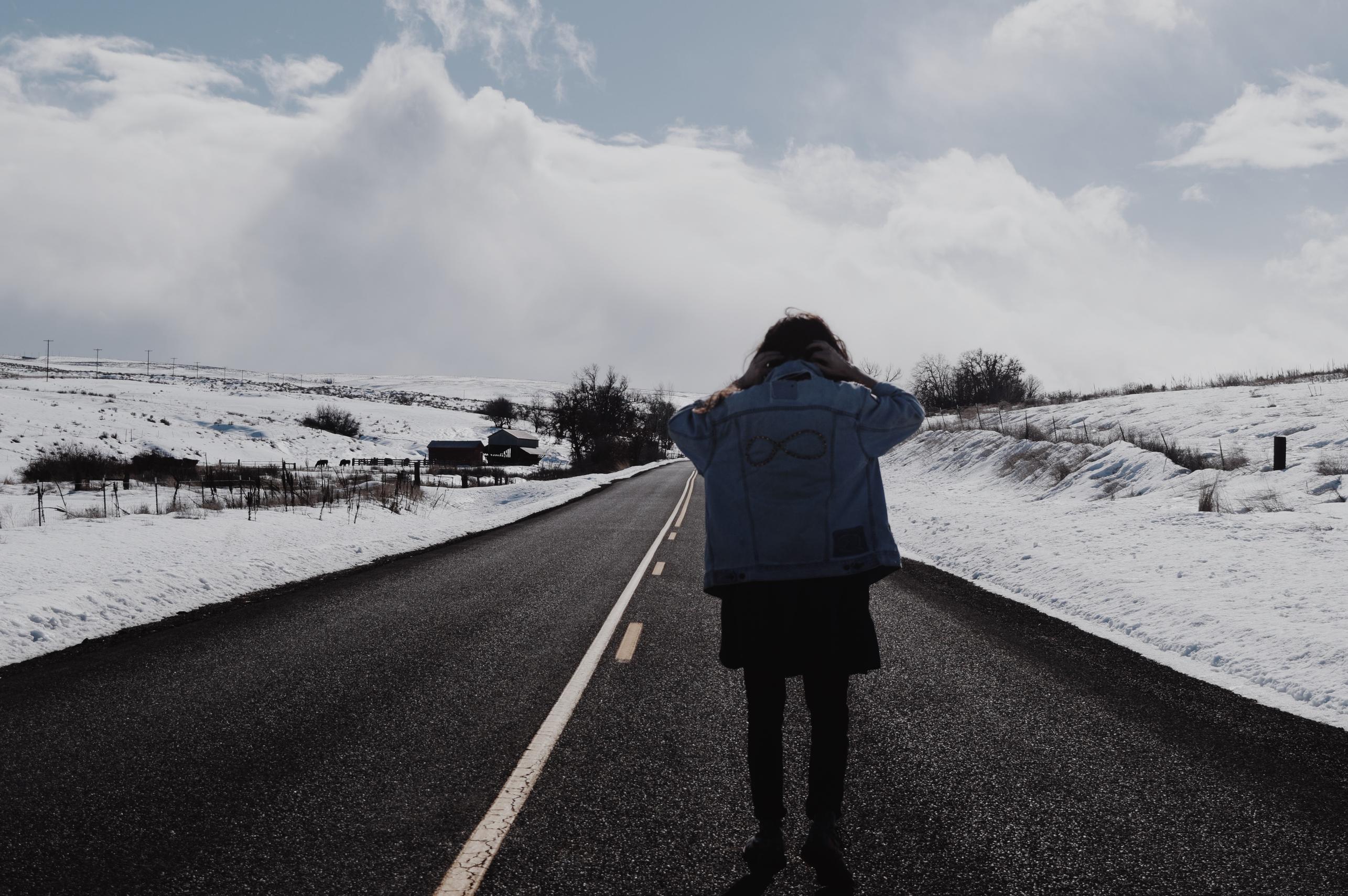 A longhaired man wearing a denim-jacket standing in the the middle of the road with his back turned. There are snow on the sides of the road, and a big cloudy sky