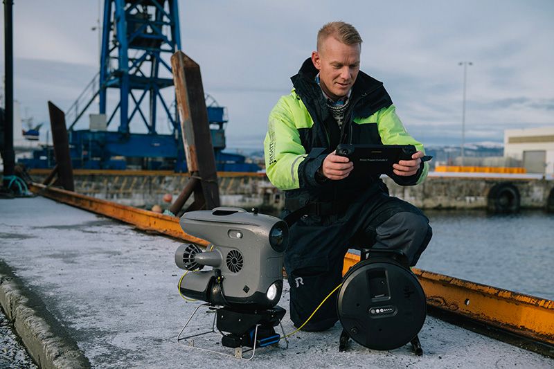 A man crouching at the docks, holding a controller, with a robot next to him