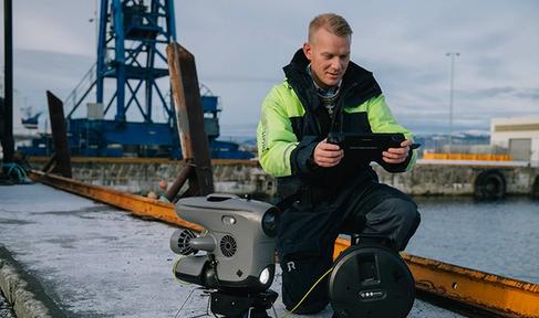 A man crouching at the docks, holding a controller, with a robot next to him