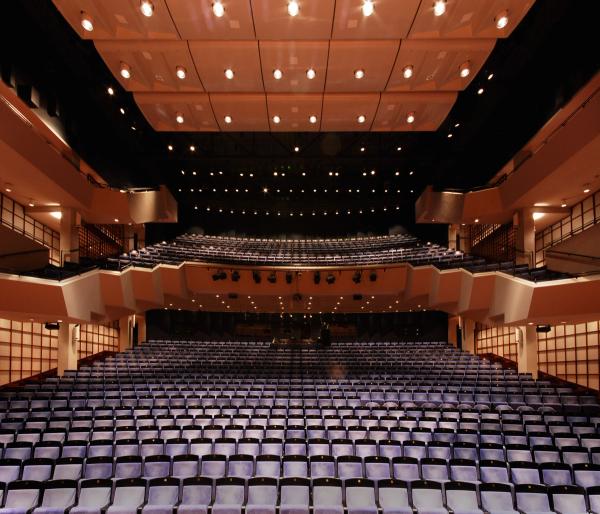 A wide shot of the interior of a modern theater, showcasing several rows of seats arranged in a semi-circular formation. The lighting is warm and even, with numerous lights mounted in the ceiling and along the walls. The architecture features a sleek, contemporary design, with angular shapes and smooth lines. The venue appears ready for a performance, with the empty seats suggesting a pre-event or rehearsal atmosphere.