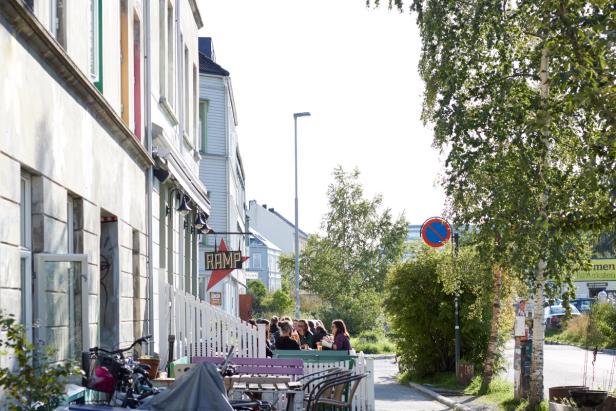 A street scene with a café or restaurant, named "RAMP," visible in the foreground. It looks like people are seated outside, likely enjoying a sunny day. The location is surrounded by greenery, with some trees and bicycle parking visible. The street appears to be relatively quiet, and there's a no-parking sign in the background, indicating a well-organized urban area.