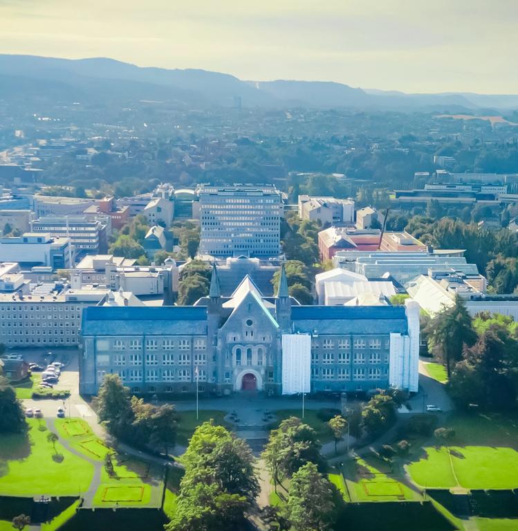 Aerial view of the NTNU Main Building in Trondheim, Norway, surrounded by lush green parks with the cityscape in the background.