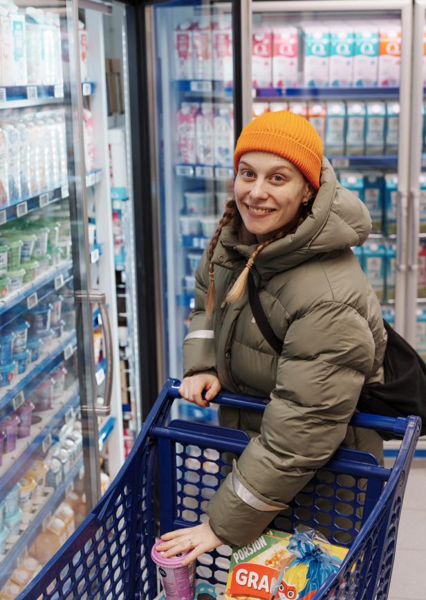 A woman in a puffy jacket and orange beanie smiles while pushing a shopping cart in a store aisle filled with dairy products.