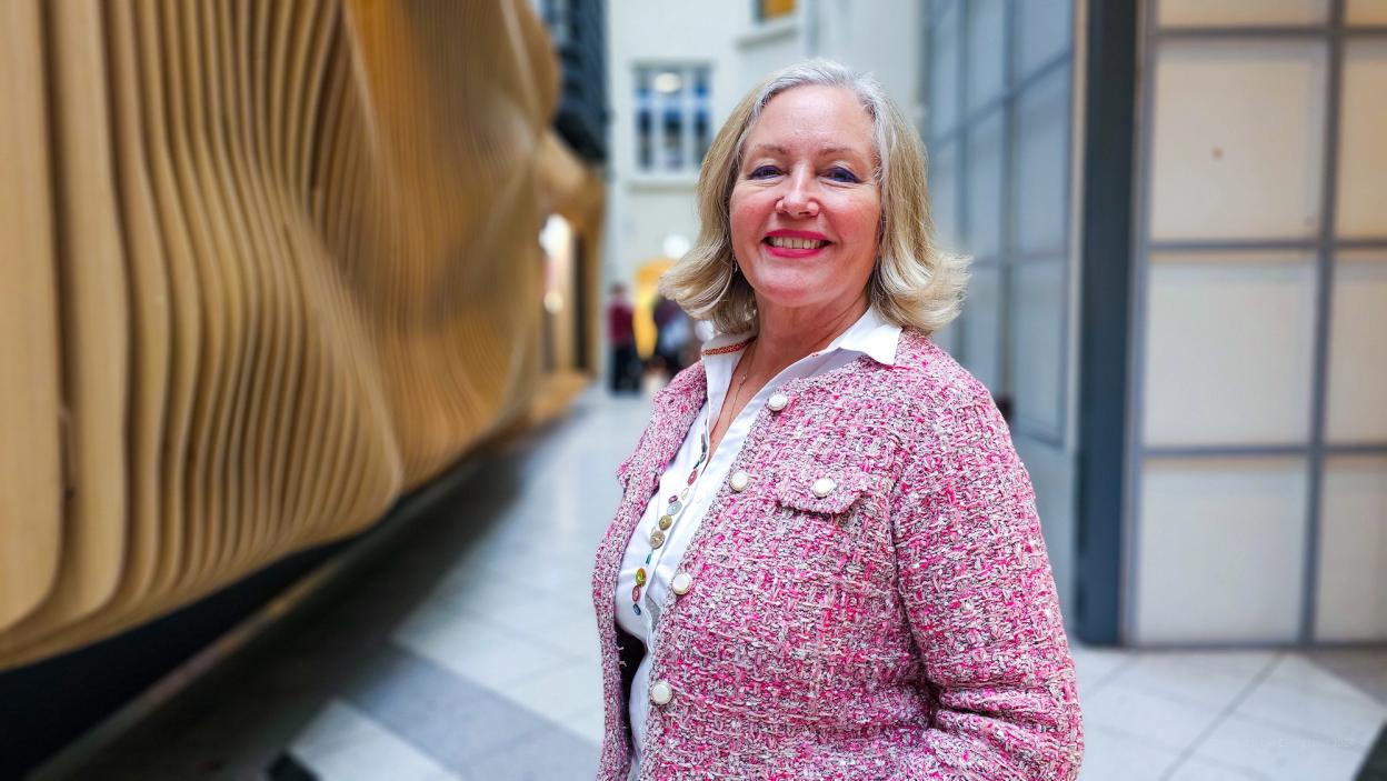 A woman in a pink tweed jacket smiling while standing in front of a modern, curved wooden installation in an indoor public space.