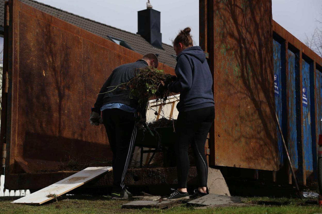 Two people lift and dispose of garden waste into a large rusty dumpster during a community cleanup event.