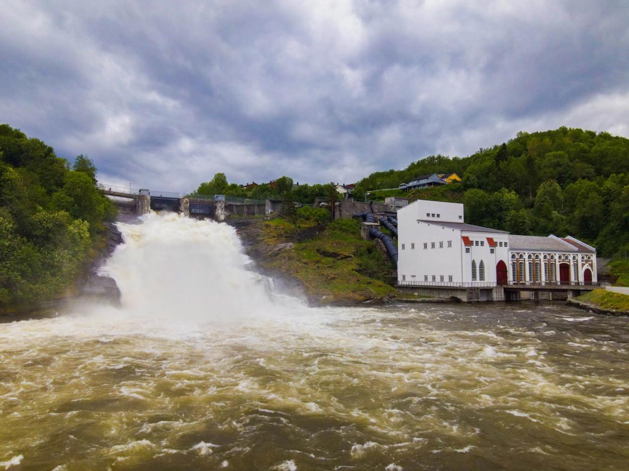 This aerial view shows the scenic Øvre Leirfoss area, with lush greenery surrounding a river and a waterfall. A hydroelectric plant is visible near the river, and walking paths run along the water, blending nature with industrial infrastructure. In the background, you can see the peaceful hills and residential areas of the region.