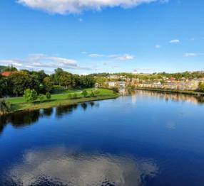 **Alt text:** A scenic view of Trondheim featuring the Nidelva River, lush greenery, and the iconic spire of Nidaros Cathedral under a clear blue sky.