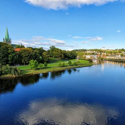 **Alt text:** A scenic view of Trondheim featuring the Nidelva River, lush greenery, and the iconic spire of Nidaros Cathedral under a clear blue sky.
