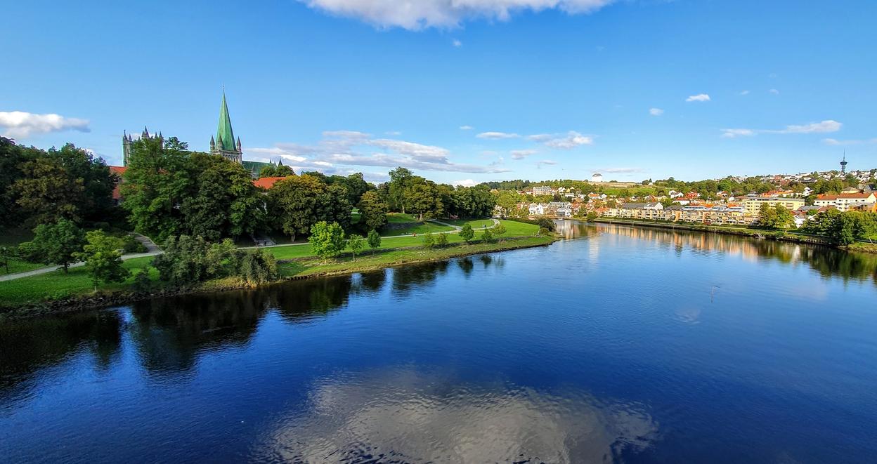 **Alt text:** A scenic view of Trondheim featuring the Nidelva River, lush greenery, and the iconic spire of Nidaros Cathedral under a clear blue sky.