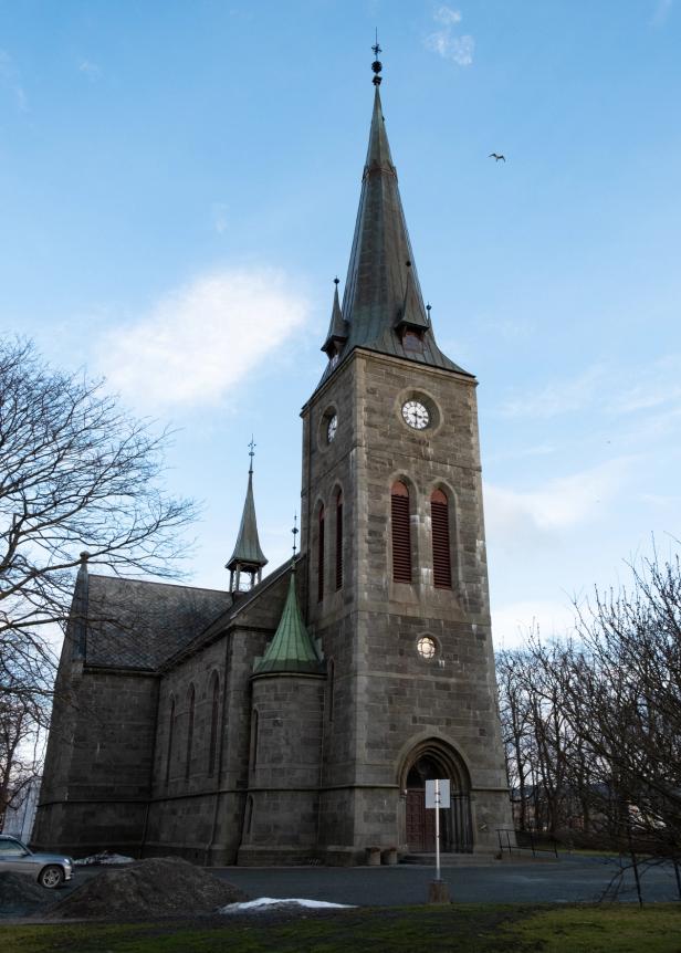 A historic stone church with a tall, pointed clock tower stands against a partly cloudy blue sky. The Gothic-style architecture features arched windows, a secondary smaller spire, and a green copper-roofed turret. Leafless trees surround the church, suggesting late autumn or early winter. A lone bird soars in the sky, adding to the serene and solemn atmosphere of the scene.