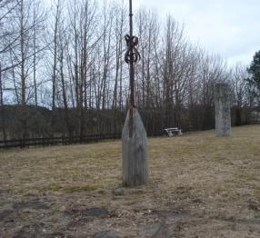The image shows a wooden church steeple or bell post next to a large stone monument in an open field. Behind the monument, there are leafless trees and a bench in the background.