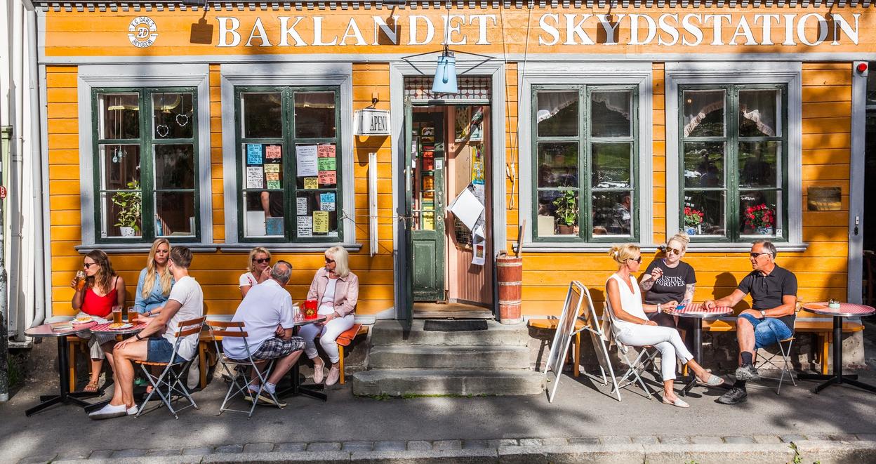 People hanging out outside of a restaurant on a beautiful summer day. You can see the shades from the trees close by on the restaurants yellow walls