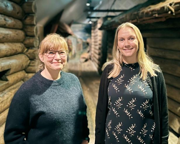 Two woman standing in front of a reconstruction of an old viking village. The houses are made of lumber, with sod roofs.