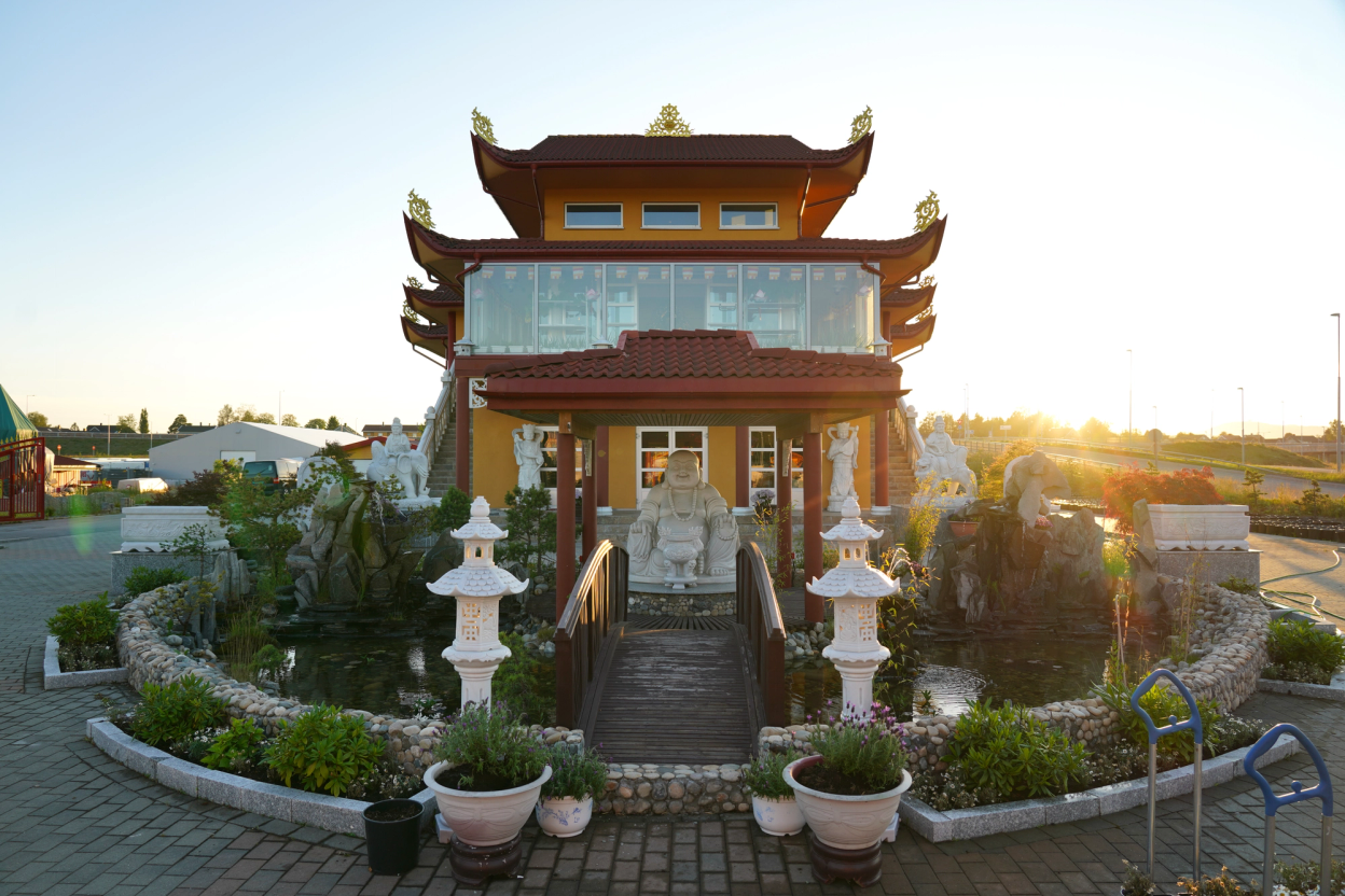 A red and yellow temple with a blue sky in the background. The sun is setting and it's a lovely day. There is a statue of Buddha in front of the temple.