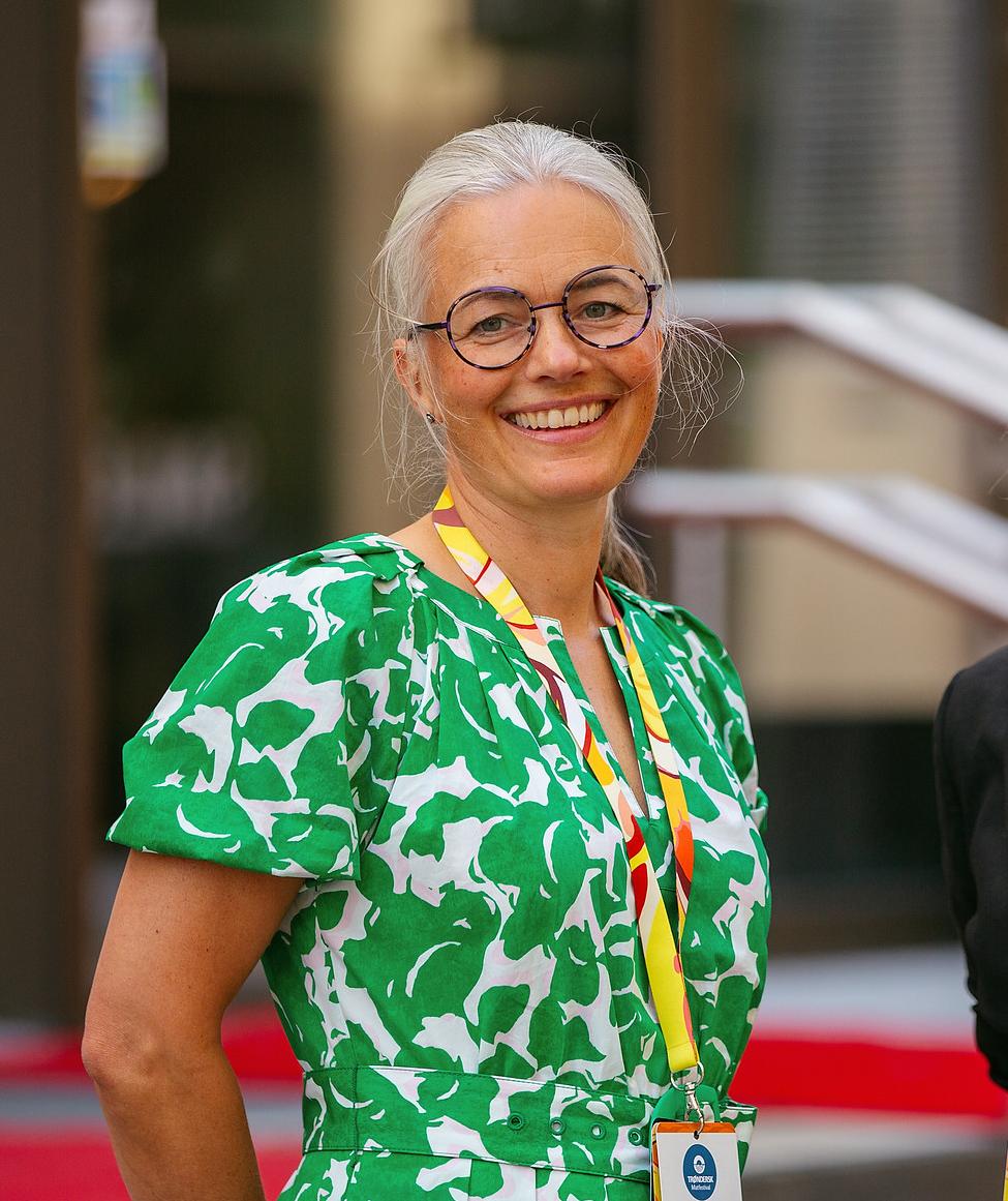 Aslaug Rustad smiling at an event, wearing a green and white patterned dress with a yellow and red lanyard holding a name badge. Her silver hair is pulled back, and she wears round glasses. The background features red carpeted steps and a modern building entrance.