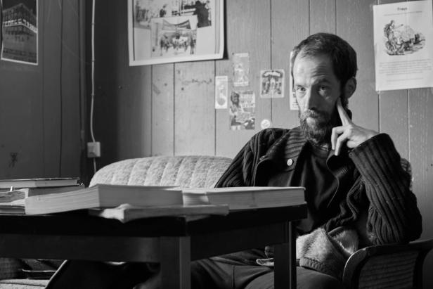 a man sitting at a table with a thoughtful expression. He's dressed in dark clothing with a long sleeve and a cardigan, looking directly at the camera. Several books are placed in front of him, and behind him is a room with wooden paneling on the walls. There are also a few posters and papers on the wall, adding to the setting's character.