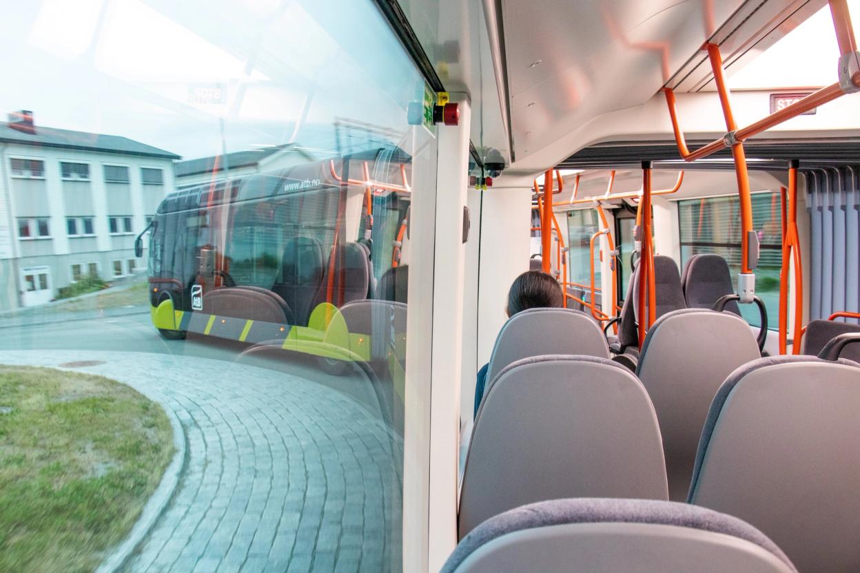 This image shows the interior of a modern metro bus, with orange handles and seats. The reflection of the bus’s exterior can be seen through the large window, along with a view of the street outside, indicating the bus is in motion. The bus seems to have a clean and sleek design, with a passenger visible in the seat.