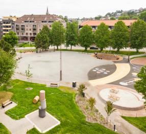 A spacious urban park with a large open gravel area, surrounded by trees and modern playgrounds featuring colorful rubber surfaces, climbing structures, and painted games. In the foreground, a small grassy section has a unique stacked sculpture and benches. Residential buildings with red and beige facades line the background, blending greenery with city life.
