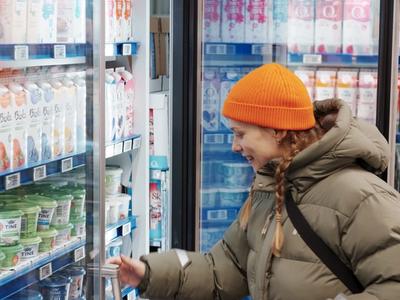 A person wearing a beige puffer jacket and bright orange beanie smiles while reaching for a product in a refrigerated section of a store. The shelves are filled with various dairy products and drinks.