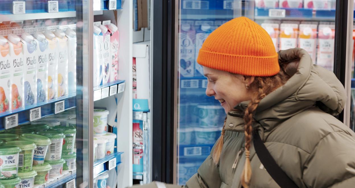 A person wearing a beige puffer jacket and bright orange beanie smiles while reaching for a product in a refrigerated section of a store. The shelves are filled with various dairy products and drinks.