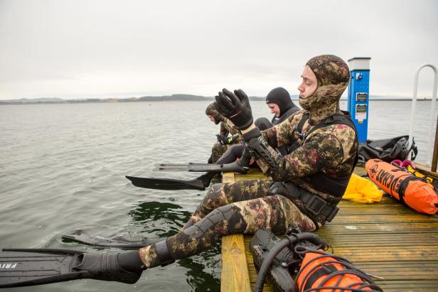 A group of free-divers sitting at the docks, getting ready to hit the sea. They are wearing camouflage-patterned diving suits