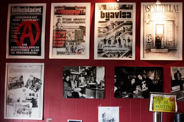 a wall decorated with historical posters and photographs. The posters appear to be from past political and social movements, with titles such as Arbeidermakt (which translates to "Workers' Power") and Byavisa (a name that likely refers to a local newspaper). The photographs on the bottom half depict people, possibly in a workplace or social setting, reflecting moments in history. The yellow sign at the bottom says, "Ingen røyte på oohkafeen," which could mean "No smoking at the café."