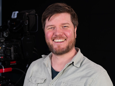 This is a photo of a person smiling, standing in front of a camera setup, possibly in a studio setting.