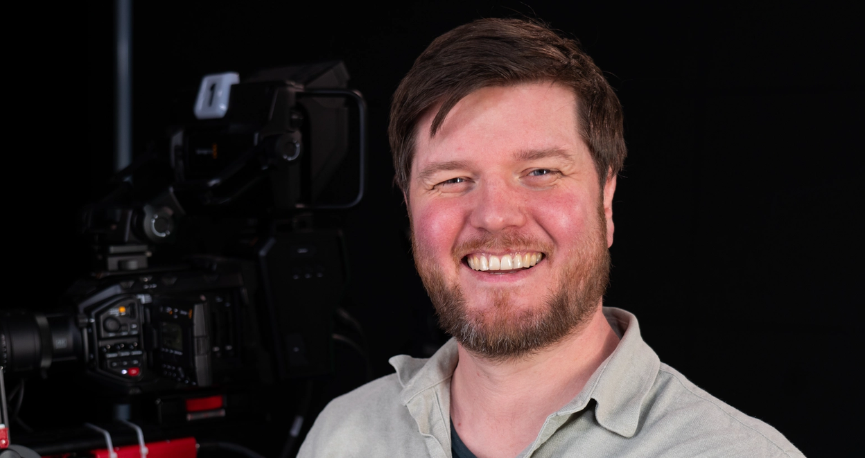 This is a photo of a person smiling, standing in front of a camera setup, possibly in a studio setting.