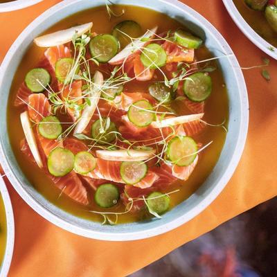 A vibrant salmon dish served at Trøndersk Matfestival. The dish features fresh slices of salmon in a golden broth, garnished with thinly sliced cucumber, apple wedges, and delicate microgreens. The bowls are placed on an orange tablecloth, adding warmth to the presentation.