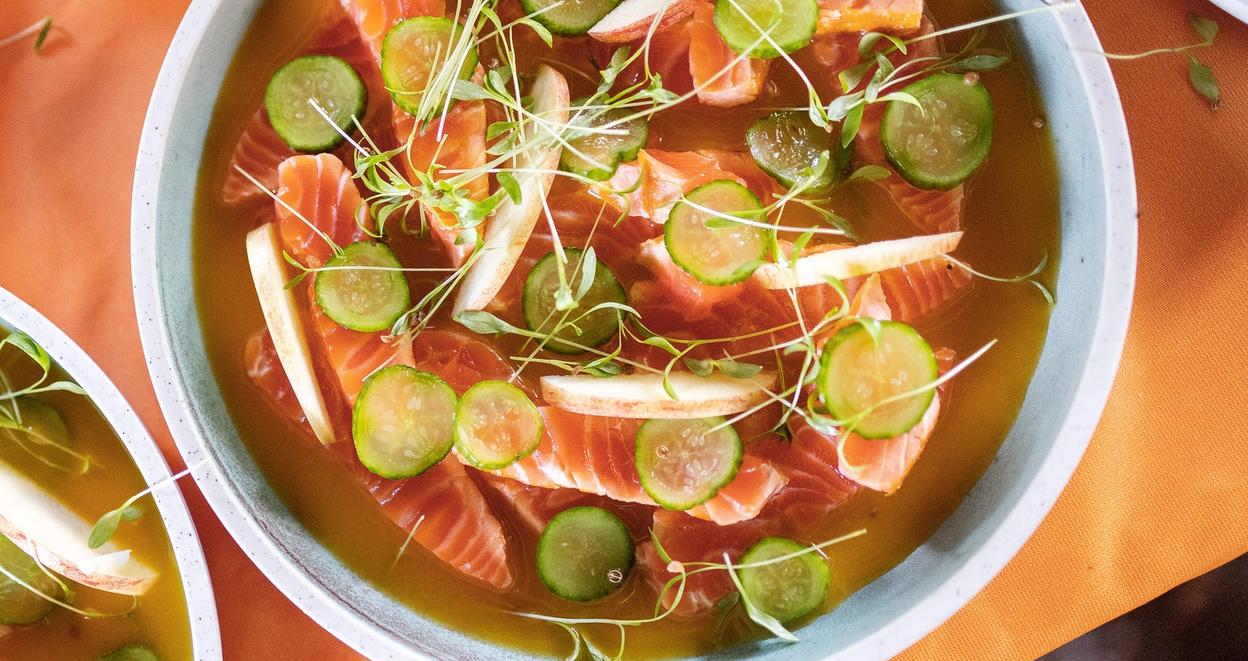 A vibrant salmon dish served at Trøndersk Matfestival. The dish features fresh slices of salmon in a golden broth, garnished with thinly sliced cucumber, apple wedges, and delicate microgreens. The bowls are placed on an orange tablecloth, adding warmth to the presentation.