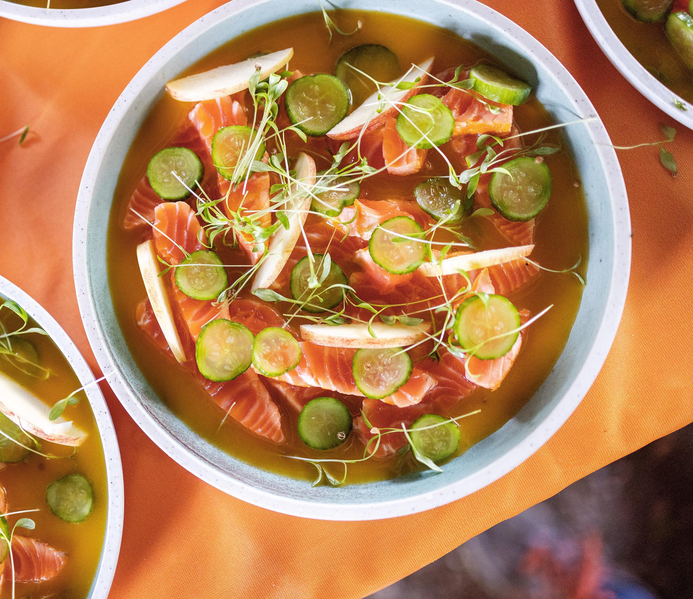 A vibrant salmon dish served at Trøndersk Matfestival. The dish features fresh slices of salmon in a golden broth, garnished with thinly sliced cucumber, apple wedges, and delicate microgreens. The bowls are placed on an orange tablecloth, adding warmth to the presentation.