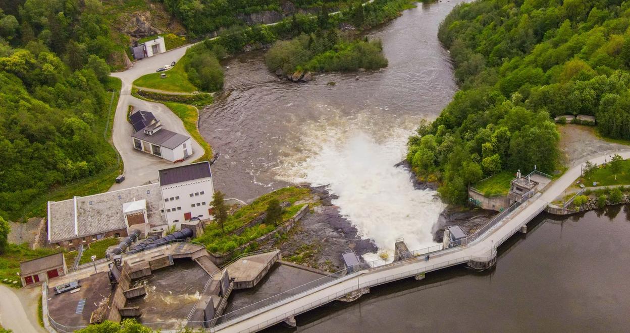 This aerial view shows the scenic Øvre Leirfoss area, with lush greenery surrounding a river and a waterfall. A hydroelectric plant is visible near the river, and walking paths run along the water, blending nature with industrial infrastructure. In the background, you can see the peaceful hills and residential areas of the region.