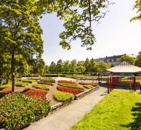 A vibrant park with neatly arranged flower beds in red, yellow, and green hues, bordered by stone edges. A winding gravel path runs through the park, shaded by lush green trees. To the right, a gazebo with a metal roof and red pillars stands near a small playground. In the background, a row of trees and buildings with dark rooftops can be seen under a bright blue sky.*