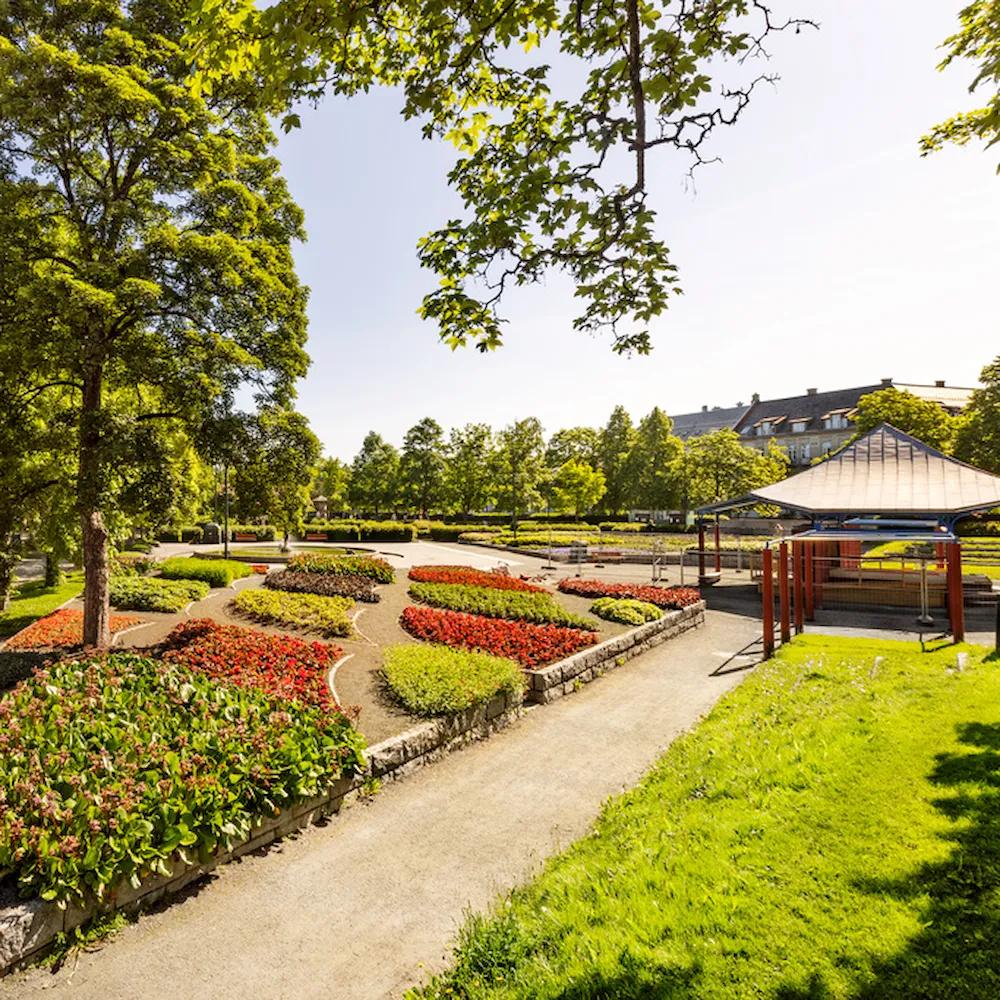 A vibrant park with neatly arranged flower beds in red, yellow, and green hues, bordered by stone edges. A winding gravel path runs through the park, shaded by lush green trees. To the right, a gazebo with a metal roof and red pillars stands near a small playground. In the background, a row of trees and buildings with dark rooftops can be seen under a bright blue sky.*