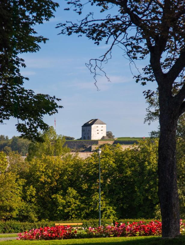 A scenic view of Kristiansten Fortress in Trondheim, Norway, framed by tree branches and lush greenery. The white, square-shaped fortress stands on a hill in the background, with a Norwegian flag flying nearby. In the foreground, a well-maintained park with a wooden bench, vibrant red flowers, and a lamppost is visible. The trees cast dappled shadows, and the sky is clear blue, indicating a bright and sunny day.