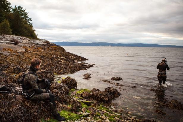 A man sitting at land watching a woman standing in the sea. She is wearing a snorkel