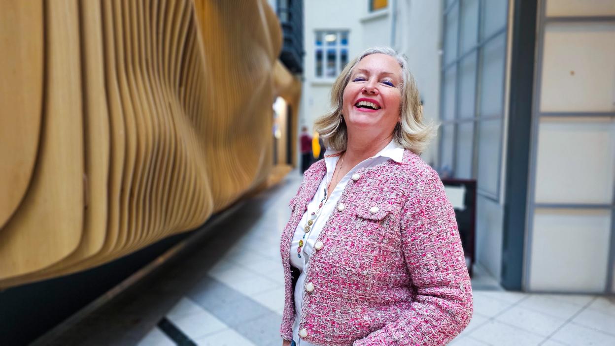 A woman in a pink tweed jacket laughing joyfully while standing in front of a modern, curved wooden installation in an indoor public space.