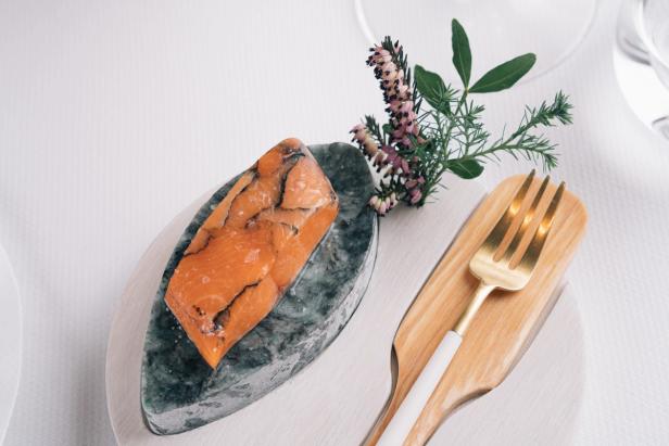 This image features an elegantly presented dish, which includes a piece of what appears to be salmon, served on a smooth stone platter. The dish is garnished with fresh herbs, and the plating style is sophisticated, with clean lines and a minimalist design. The utensils, including a golden fork and a wooden-handled knife, complement the refined aesthetic of the dish.