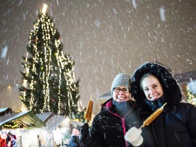 Two people enjoying holiday treats at a snowy Christmas market with a beautifully lit tree in the background.