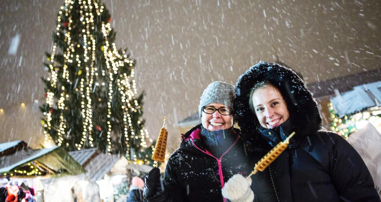 Two people enjoying holiday treats at a snowy Christmas market with a beautifully lit tree in the background.