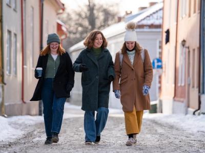 Three young women dressed in warm winter clothing walk together on a snowy cobblestone street, smiling and chatting. One holds a coffee cup, and they are surrounded by colorful historic buildings in a charming winter setting.