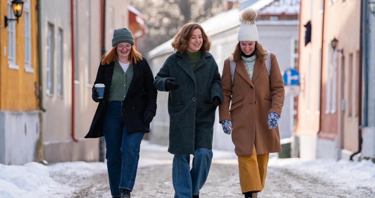 Three young women dressed in warm winter clothing walk together on a snowy cobblestone street, smiling and chatting. One holds a coffee cup, and they are surrounded by colorful historic buildings in a charming winter setting.
