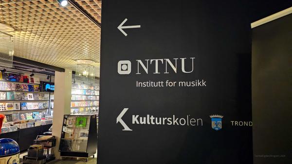 A black sign inside a building, directing visitors to the NTNU Institute of Music and the Kulturskolen (Cultural School) in Trondheim. A record store is visible in the background.