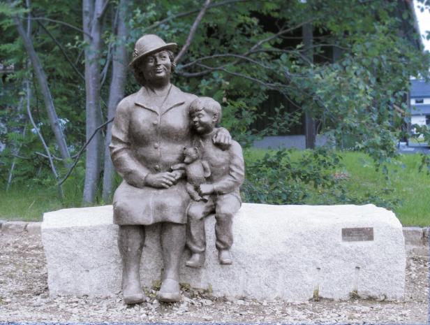 A metal statue of a woman sitting with her child on a stone bench. There's a metal plaque on the bench with the artist's details.