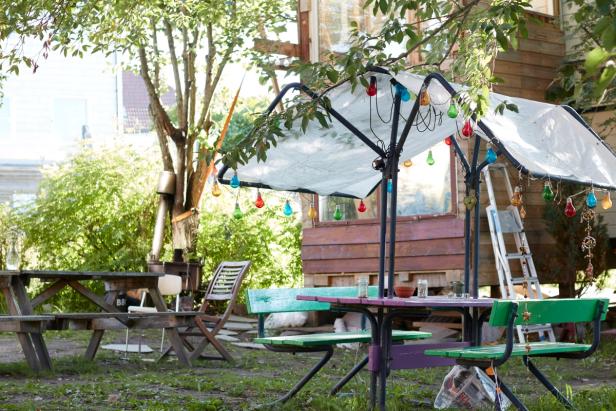 a cozy outdoor setting with colorful string lights hanging from a tree, adding a vibrant and playful touch to the space. There are two green tables with chairs, and a wooden bench in the background. The area is surrounded by greenery, including plants and trees, and the buildings in the background suggest it is part of a community or social gathering space.