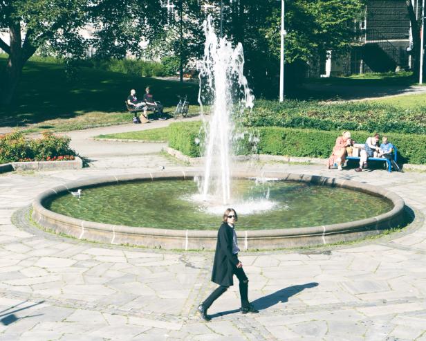 A mysterious black-clad man wearing sunglasses walking in front of a big fountain. It's a nice summer day, and there's a family enjoying some lunch on a bench in the background